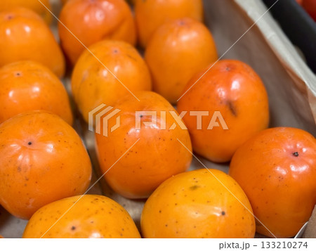 A pile of ripe juicy orange persimmons in a box, on sale at a supermarket vegetable stand, showcases organic, vegetarian and healthy food. Close-up. 133210274