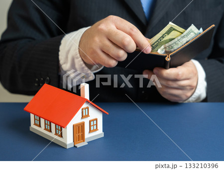 A businessman counts money near a suburban house. Household bills and maintenance. Monthly mortgage payments. family budgeting, rising living costs, finance planning for real estate. 133210396