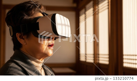Asian elderly woman wearing virtual reality headset experiencing VR technology in warm sunlit traditional wooden interior room 133210774