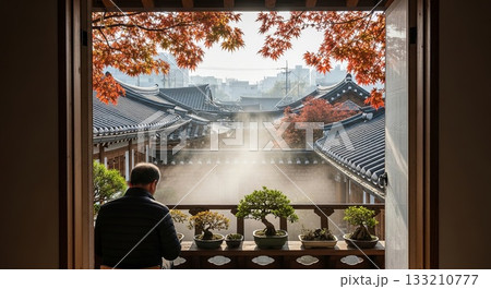 Asian man contemplating traditional Korean hanok architecture with bonsai trees on windowsill during autumn season with morning mist 133210777