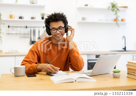 A young man wearing headphones and glasses smiles while taking notes at a table with a laptop and a cup of coffee. A young man wearing headphones and glasses smiles while taking notes at a table with a laptop and a cup of coffee. 133211339