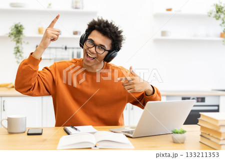 A young man with headphones smiles and points while working on a laptop at home. 133211353
