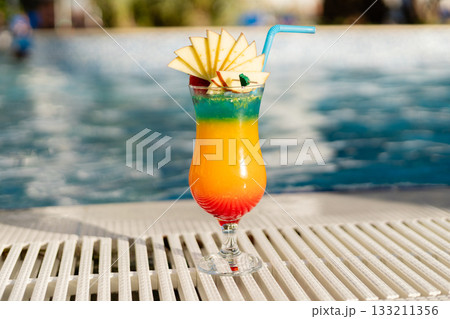 A vibrant cocktail with layers of orange, green, and red sits beside a pool. The drink is garnished with fresh apple slices and a cherry, showcasing a refreshing vibe 133211356