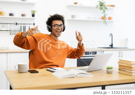 A joyful man with headphones points at the camera while working on a laptop at home. 133211357