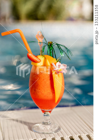 A vibrant orange cocktail sits beside a refreshing pool, adorned with fruit slices and colorful decorations. Sunlight sparkles on the water, creating a relaxing atmosphere 133211358
