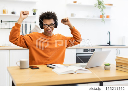 An enthusiastic young man celebrates a success while working on his laptop at home, wearing headphones and glasses. An enthusiastic young man celebrates a success while working on his laptop at home, wearing headphones and glasses. 133211362