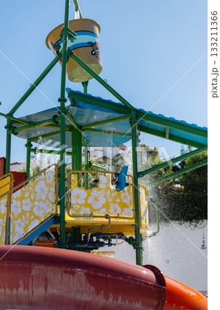 Bright water play area features a large splash tower with water cascading down. Nearby, colorful slides invite children to enjoy a fun day in the sun during summer 133211366