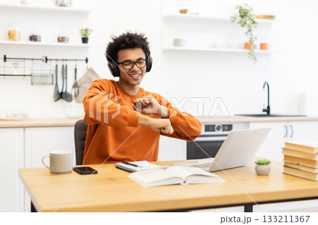 A young man with headphones dances joyfully while working on his laptop at home, enjoying music and his work. A young man with headphones dances joyfully while working on his laptop at home, enjoying music and his work. 133211367