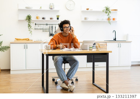 A young man with headphones smiles while working on a laptop at a table in his kitchen. A young man with headphones smiles while working on a laptop at a table in his kitchen. 133211397