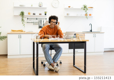 A young man with headphones works on his laptop at a table in his kitchen, smiling while taking notes. A young man with headphones works on his laptop at a table in his kitchen, smiling while taking notes. 133211413