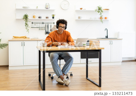 A young man with glasses and headphones works on a laptop at a table in his kitchen, taking notes. A young man with glasses and headphones works on a laptop at a table in his kitchen, taking notes. 133211473
