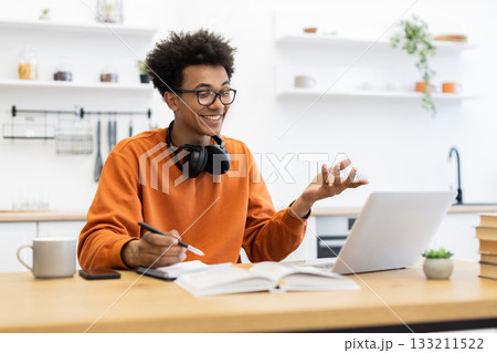 A young man with glasses smiles while participating in a video call on his laptop, gesturing with his hands. A young man with glasses smiles while participating in a video call on his laptop, gesturing with his hands. 133211522