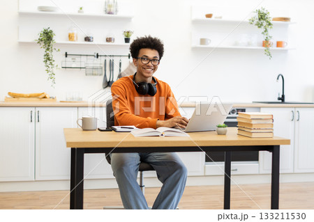A young man with glasses smiles while working on his laptop at a wooden table in a bright kitchen setting. A young man with glasses smiles while working on his laptop at a wooden table in a bright kitchen setting. 133211530