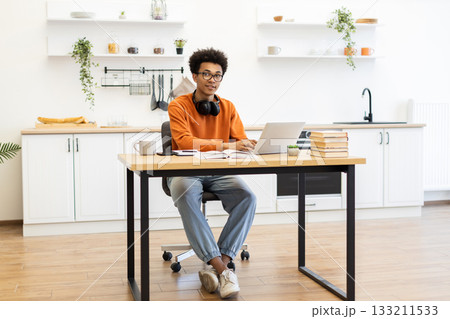 A young man with glasses sits at a desk, working on a laptop in a bright, modern kitchen setting. A young man with glasses sits at a desk, working on a laptop in a bright, modern kitchen setting. 133211533