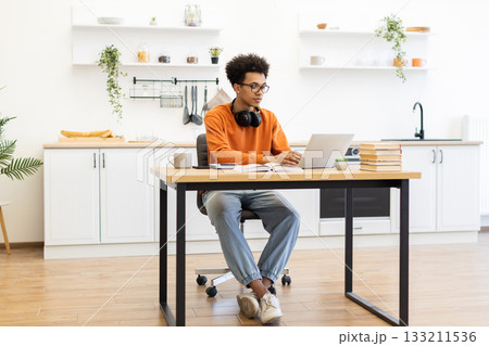 A young man with glasses works on a laptop at a desk in a modern kitchen setting, with headphones around his neck. A young man with glasses works on a laptop at a desk in a modern kitchen setting, with headphones around his neck. 133211536