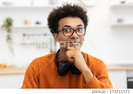 A young Black man with glasses smiles, resting his chin on his hand, with headphones around his neck. A young Black man with glasses smiles, resting his chin on his hand, with headphones around his neck. 133211572