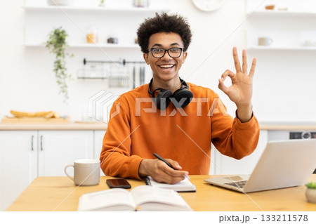 A cheerful young man with glasses smiles and makes an okay gesture while working at a desk with a laptop and notebook. 133211578