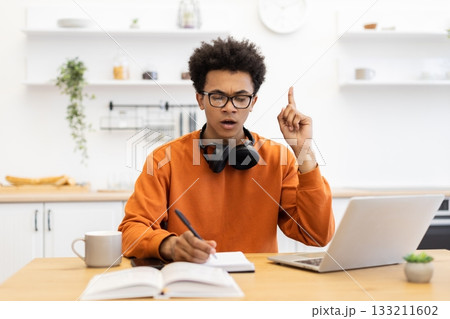 A young man with glasses is working on his laptop and writing in a notebook while having an idea. 133211602