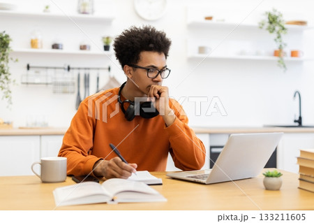 A young man with glasses is working on a laptop at home, taking notes and looking thoughtful. A young man with glasses is working on a laptop at home, taking notes and looking thoughtful. 133211605