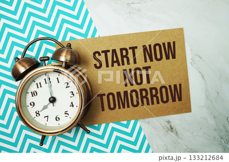 Start Now Not Tomorrow text written on paper card with alarm clock on marble background Start Now Not Tomorrow text written on paper card with alarm clock on marble background 133212684