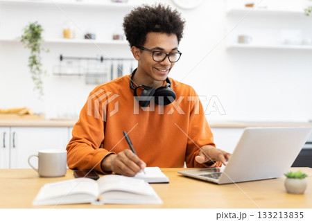A young man with glasses works on a laptop while taking notes in a bright kitchen setting. 133213835
