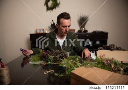 Man Crafts Christmas Wreaths With Pine Cones And Greenery At Cozy Home Table 133215515