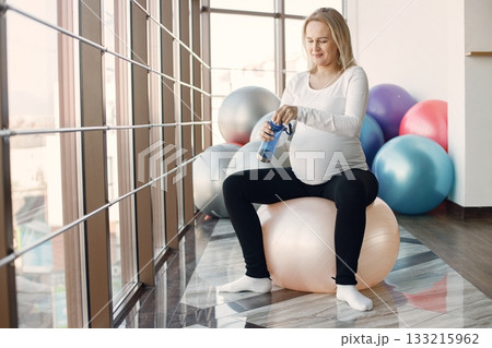 Caucasian pregnant woman in third trimester drinking water from sportive bottle. Woman in bright fitness studio with big windows. Blonde woman wearing whit shirt and black leggins. Caucasian pregnant woman in third trimester drinking water from sportive bottle. Woman in bright fitness studio with big windows. Blonde woman wearing whit shirt and black leggins. 133215962