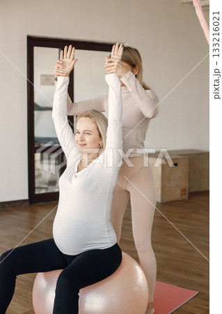 Pregnant woman doing yoga with personal trainer. Yoga trainer assisting pregnant woman while doing exercises with a fitball. Blonde pregnant woman wearing white shirt. Pregnant woman doing yoga with personal trainer. Yoga trainer assisting pregnant woman while doing exercises with a fitball. Blonde pregnant woman wearing white shirt. 133216021