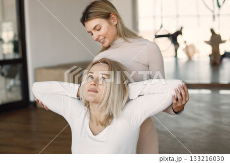 Blonde woman doing yoga with personal trainer. Yoga trainer assisting woman while doing exercises. Blonde woman wearing white clothes and trainer wearing beige costume. Blonde woman doing yoga with personal trainer. Yoga trainer assisting woman while doing exercises. Blonde woman wearing white clothes and trainer wearing beige costume. 133216030