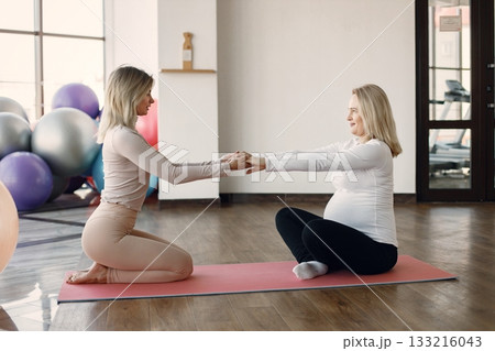 Pregnant woman doing yoga with personal trainer. Yoga trainer assisting pregnant woman while doing exercises. Blonde pregnant woman wearing black and white clothes. Pregnant woman doing yoga with personal trainer. Yoga trainer assisting pregnant woman while doing exercises. Blonde pregnant woman wearing black and white clothes. 133216043