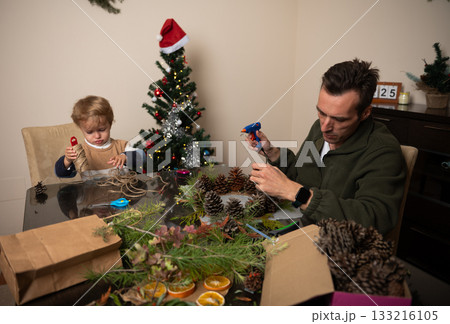 Family Christmas Craft At The Table With Pinecones, Twine, And A Decorated Tree In A Cozy Home 133216105