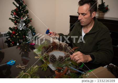 Man Crafts Christmas Wreath With Pinecones At Home For Cozy Holiday Decor Man Crafts Christmas Wreath With Pinecones At Home For Cozy Holiday Decor 133216109