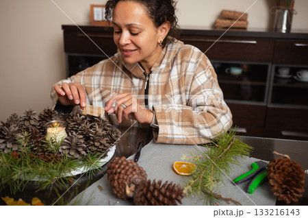 Woman Crafts Christmas Wreath With Pine Cones And Candle In Cozy Home Setting Woman Crafts Christmas Wreath With Pine Cones And Candle In Cozy Home Setting 133216143