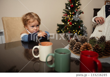Young Child At Table With Christmas Tree Decor And Pinecones, Family Moment Young Child At Table With Christmas Tree Decor And Pinecones, Family Moment 133216285