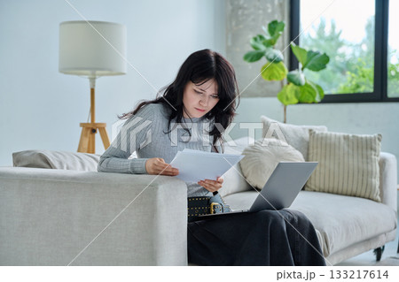 Young serious student girl sitting at home on couch with laptop, reading papers 133217614