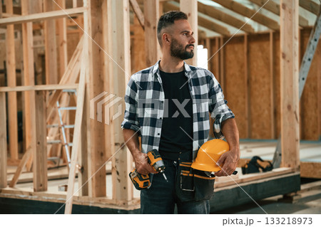 Building the house, holding hard hat and drill. Industrial worker in wooden warehouse 133218973