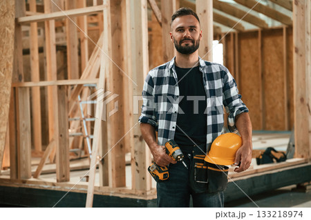 Building the house, holding hard hat and drill. Industrial worker in wooden warehouse 133218974