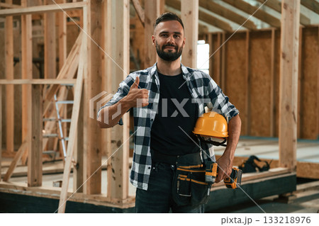 Building the house, holding hard hat and drill. Industrial worker in wooden warehouse 133218976