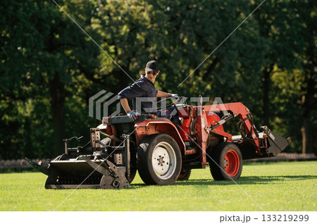 Sitting and driving. Man is with utility tractor with grass cutter and aerator equipment on the field Sitting and driving. Man is with utility tractor with grass cutter and aerator equipment on the field 133219299