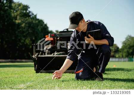 Quality control, touching the grass. Man is with utility tractor with grass cutter and aerator equipment on the field Quality control, touching the grass. Man is with utility tractor with grass cutter and aerator equipment on the field 133219305