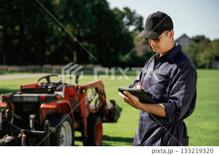 Black digital tablet in hands. Man is with utility tractor with grass cutter and aerator equipment on the field 133219320