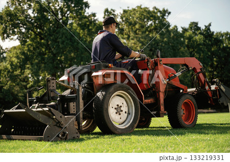Sitting and driving. Man is with utility tractor with grass cutter and aerator equipment on the field Sitting and driving. Man is with utility tractor with grass cutter and aerator equipment on the field 133219331