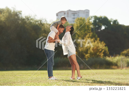 Kid is sitting on the shoulders. Mother and father are with son outdoors on the field 133221821
