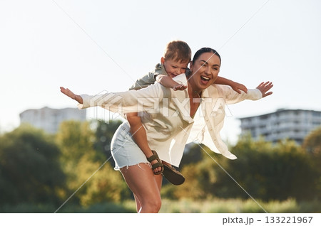 Holding kid on the shoulders. Woman with her little son are on the field together Holding kid on the shoulders. Woman with her little son are on the field together 133221967