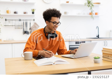 A young man with glasses works on a laptop while taking notes in a kitchen setting, smiling. 133223127