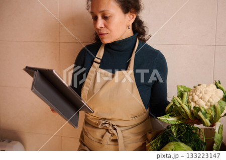 Focused Woman In Apron With Tablet In Kitchen Handling Cauliflower For Market Or Recipe Focused Woman In Apron With Tablet In Kitchen Handling Cauliflower For Market Or Recipe 133223147
