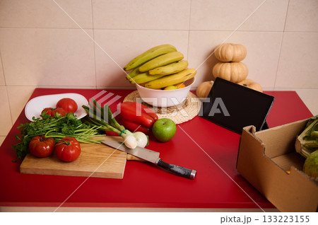 Fresh Produce on Red Kitchen Counter With Bananas, Tomatoes, Greens, and a Tablet 133223155