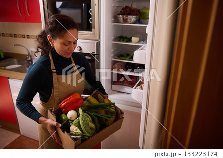 Woman With Apron Carries Box Of Fresh Groceries From Open Refrigerator In Bright Kitchen 133223174