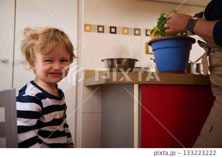Happy Toddler In Striped Shirt Smiles At Camera In Kitchen While Parent Prepares Food 133223222
