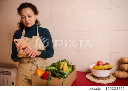 Woman In Apron Checks Fresh Produce Box On Kitchen Counter Using Smartphone For Shopping 133223251